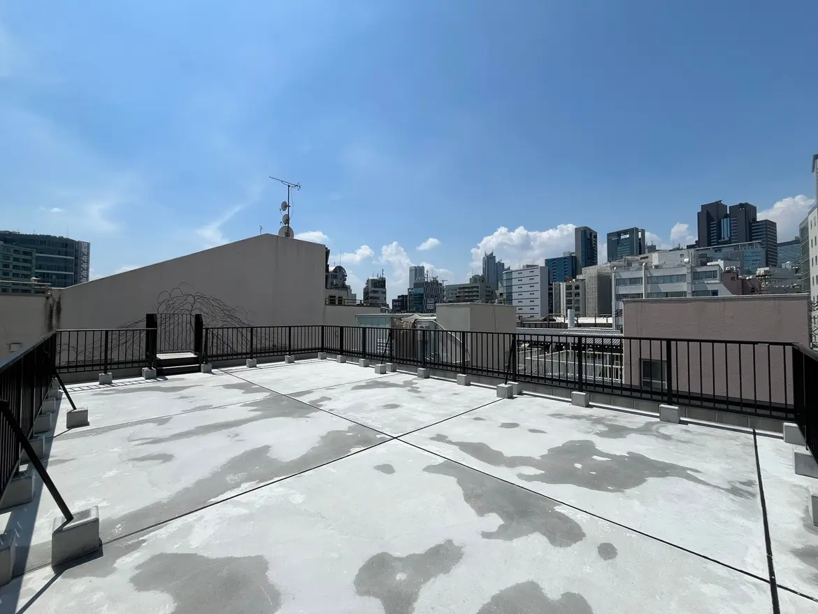 - 都会的な景色が広がる屋上テラス、青空と白い雲、金属製のフェンスとコンクリート床のあるスペース