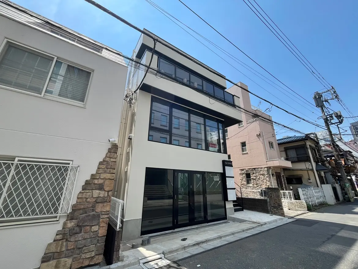 - モダンな外観の3階建て住宅、ガラスの大きな窓と白い壁、隣接する伝統的な住宅や電柱が見える街並み