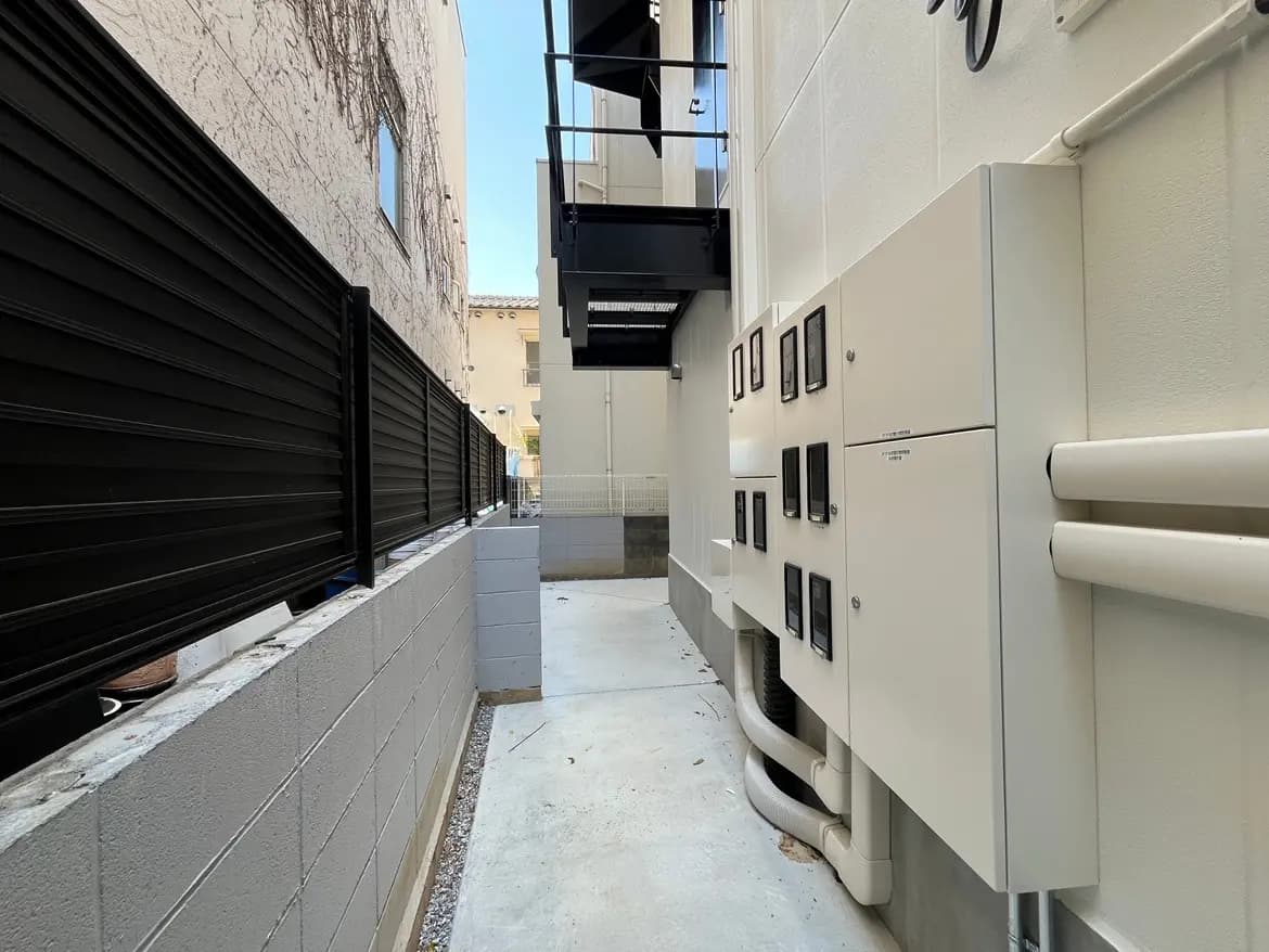 - 建物の側面に設置された電気メーターと配管、ロイヤルブルーのフェンスが並ぶ狭い通路の風景