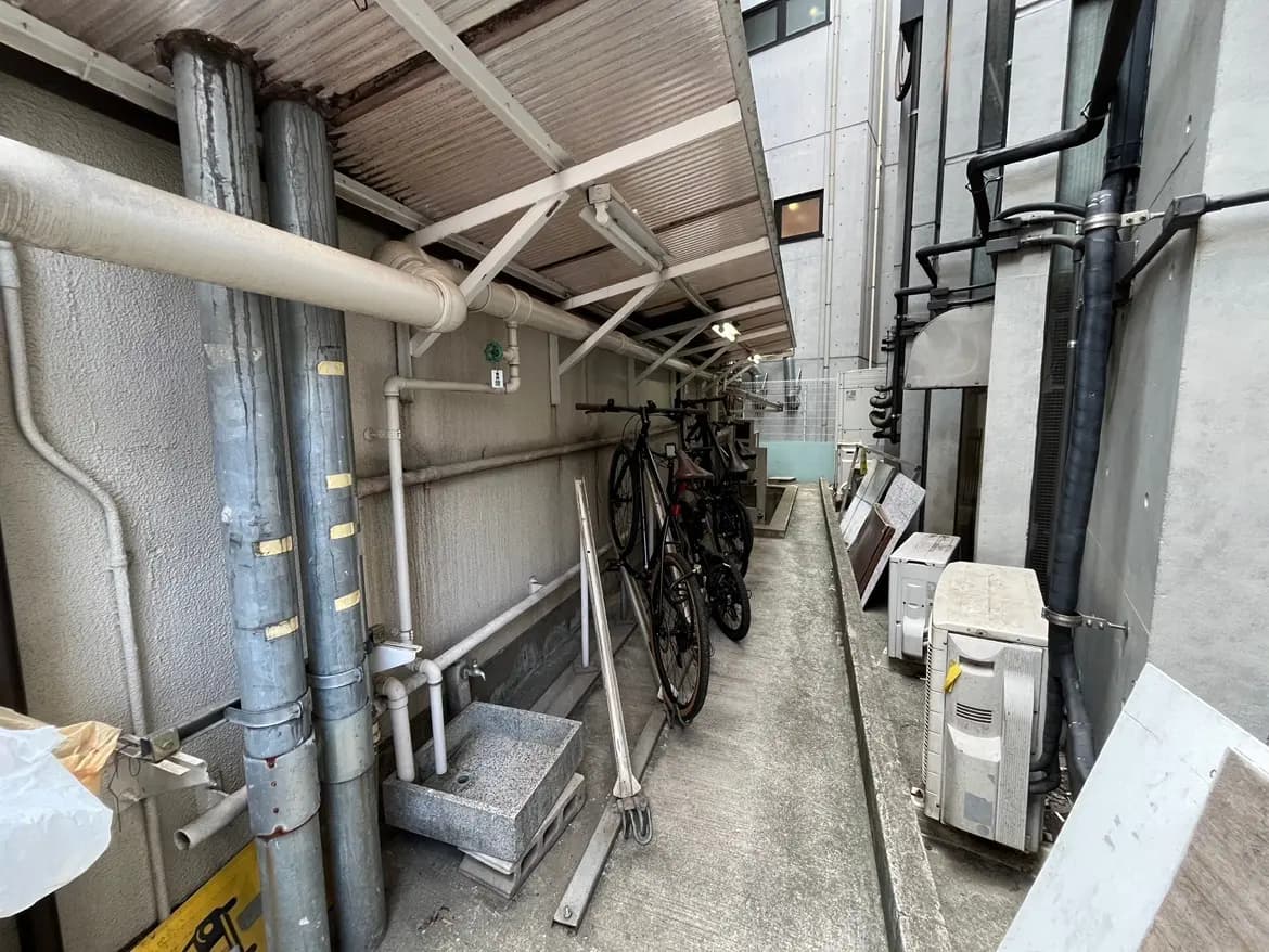 - 駐輪場の通路、壁面に配管があり、自転車が2台並んでいる狭い空間の写真