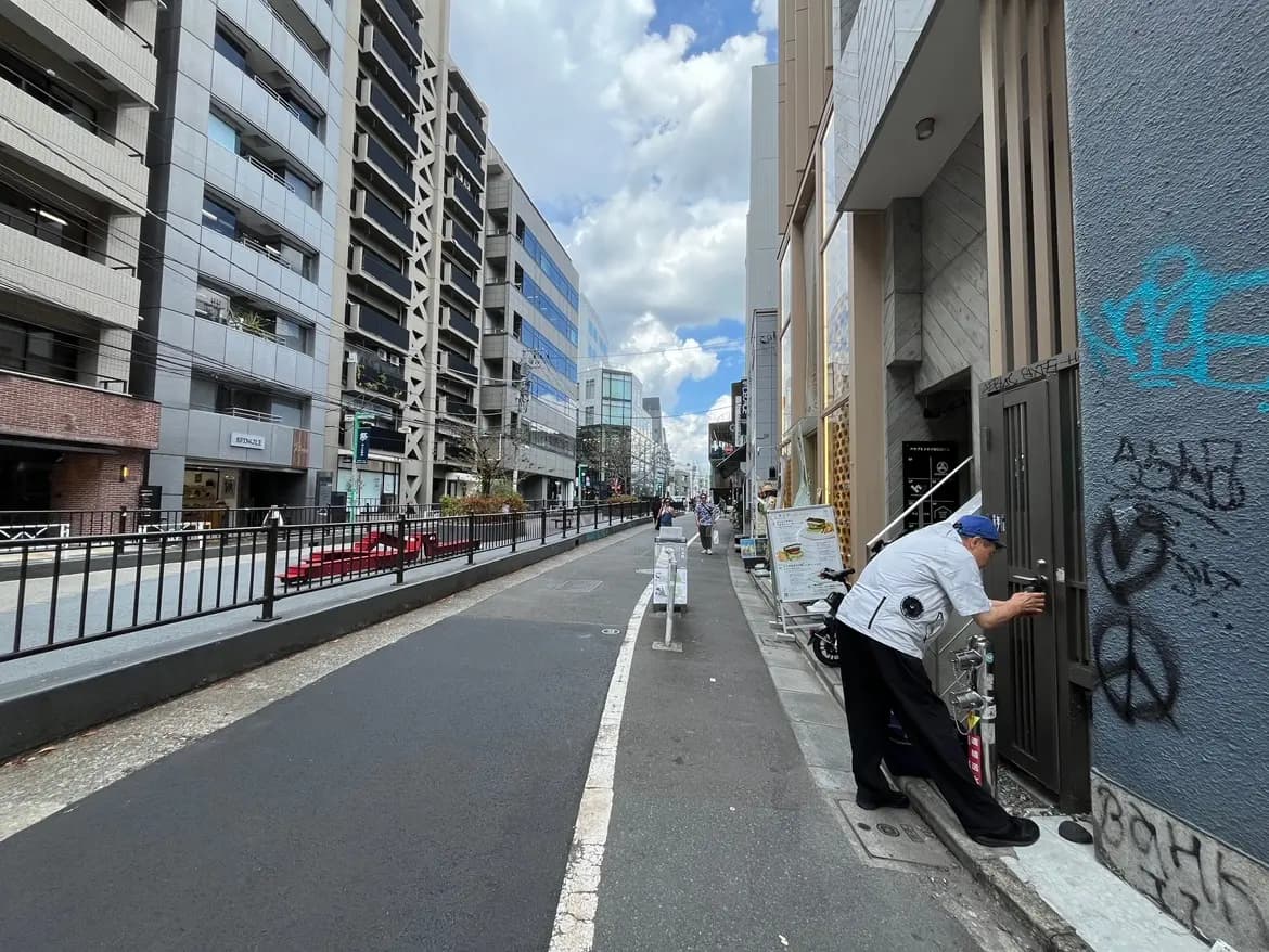 都市の商業エリアの通り、現代的な建物とグラフィティが施された壁、歩道に立つ人々の姿がある風景