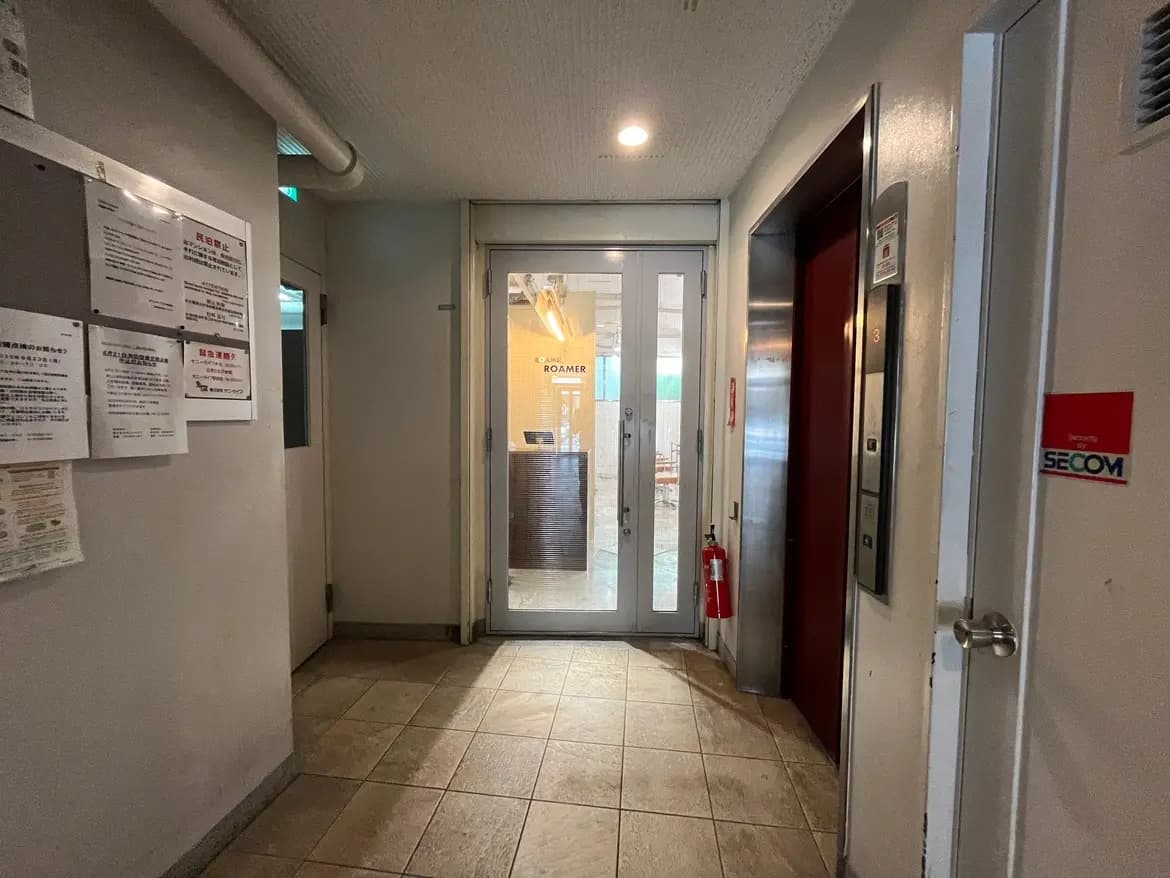 - 暗い廊下のエレベーター前、ガラスドアの先にある明るいエントランス、壁には掲示物が貼られたインテリア