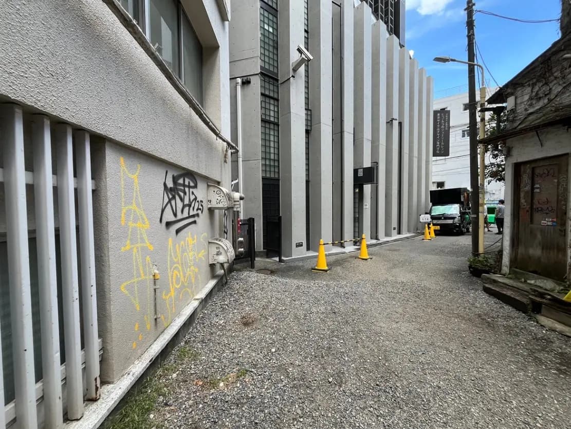 - 都市の裏通りにあるビルの側面、グラフィティが描かれた壁と砂利道、周辺に立つ交通コーンと建物の特徴的なデザイン