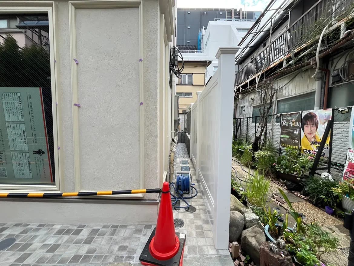 - 建物の外壁と施工作業中の小道、赤いコーンと庭の植物が見える風景