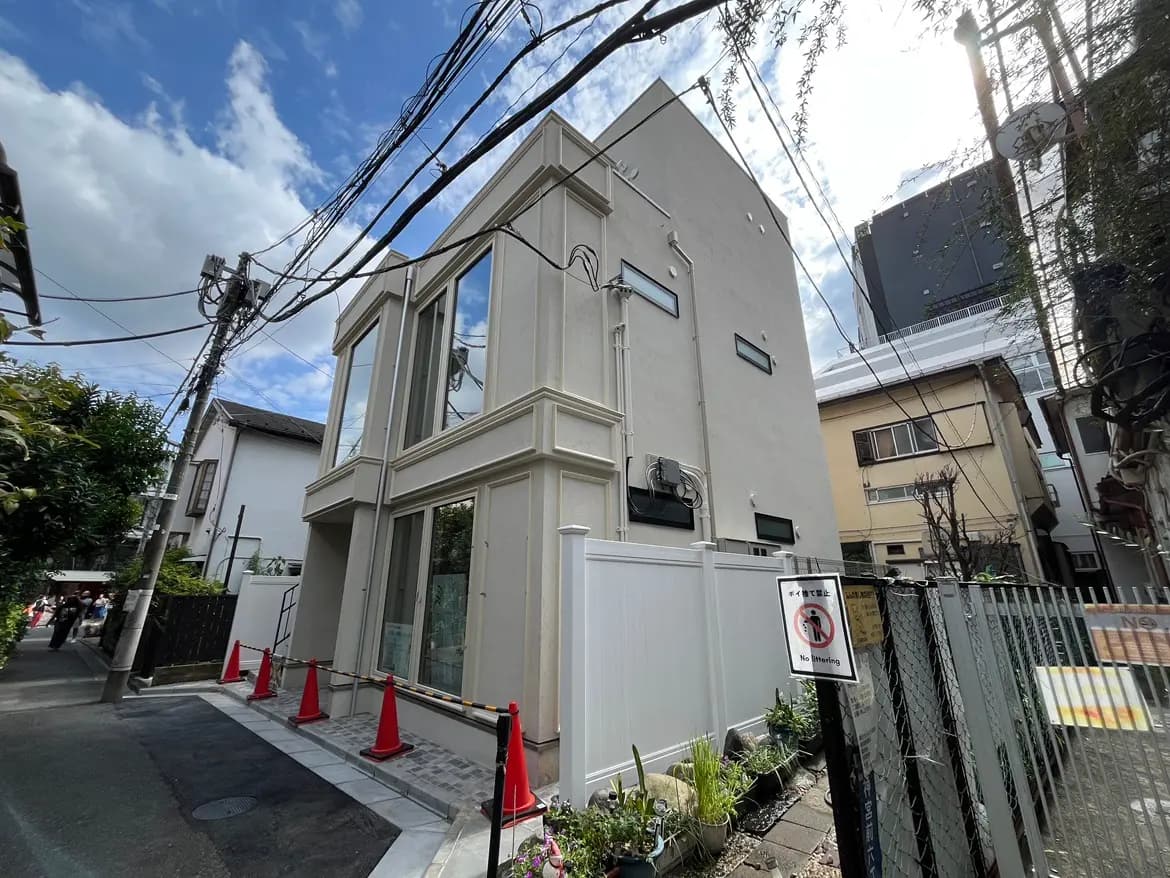 - モダンな外観の三階建て住宅、透明な窓と白い壁材、周囲には緑の植栽と交通標識がある街並み