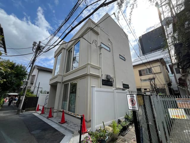 - モダンな外観の三階建て住宅、透明な窓と白い壁材、周囲には緑の植栽と交通標識がある街並み