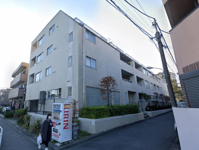 - モダンなアパートメントの外観、近くに自動販売機と緑の植栽、通りに面したシンプルなデザインの建物