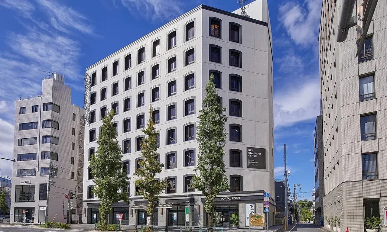 - 現代的なデザインのオフィスビル、周囲に高い木々と他の建物が並ぶ街の風景