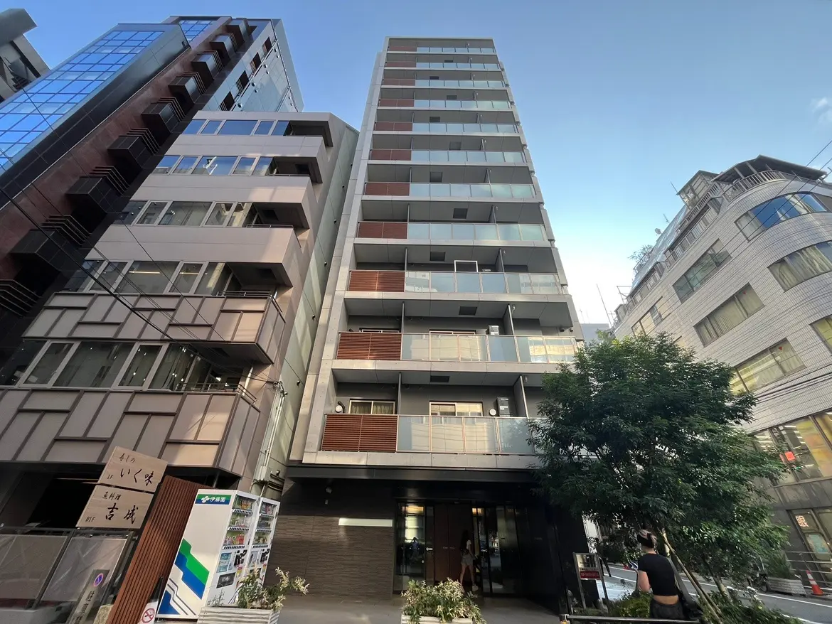 - モダンなデザインのビル外観、周囲に高層ビルが立ち並ぶ都市風景、エントランスには緑の植栽がある