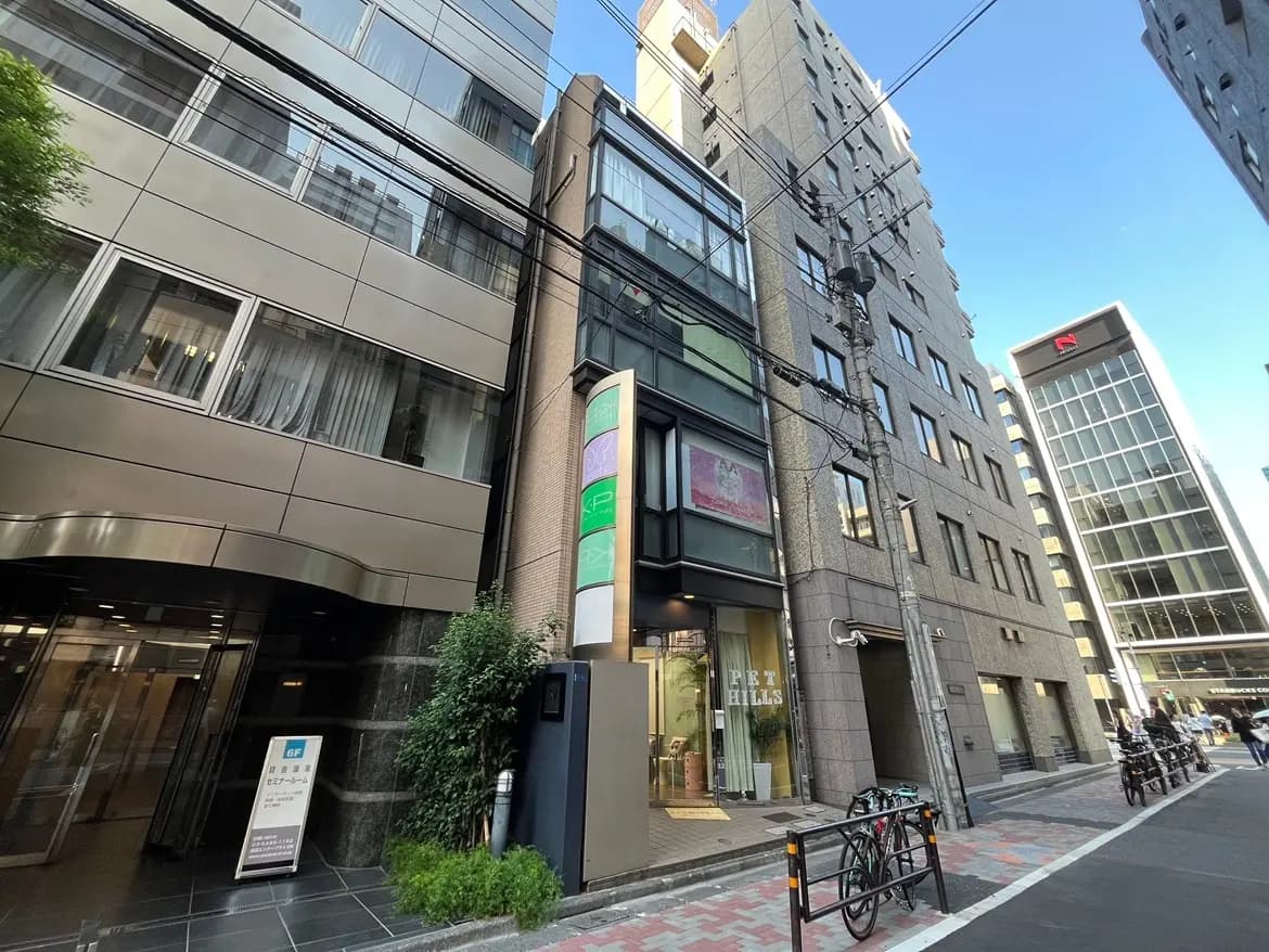 - 都会的なデザインのビル外観、高層ビルと並ぶモダンなファサード、通りに面したガラス張りのエントランスと看板