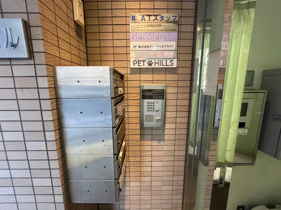 - 銀色の郵便受けが並ぶエントランス、壁にはインターホンと各種プレートが掲示された明るい雰囲気の入り口