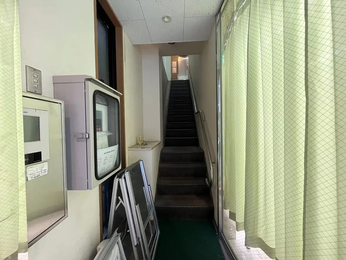 - 明るい色のカーテンが掛かった通路、左側に電気系統計器と階段が見えるシンプルなインテリア