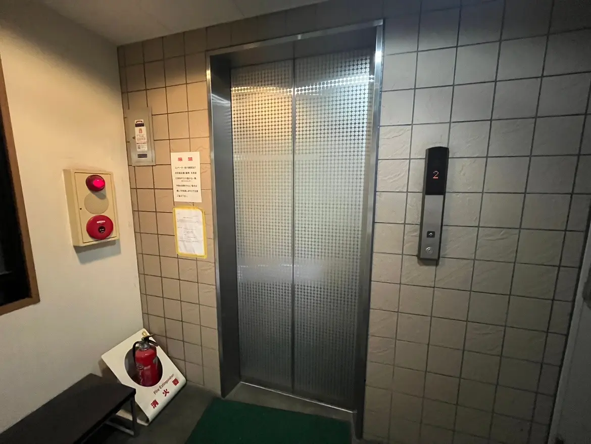 - クリーム色のタイルの壁に囲まれたエレベーターの前景、消防用の赤いベルと消火器が設置された安全対策の様子