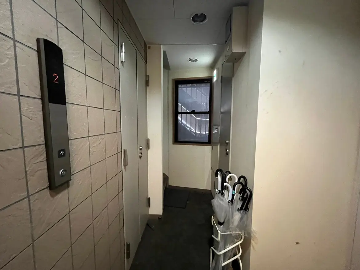 エレベーターの近くに位置する静かな廊下、バックグラウンドには雨傘を収納するスタンドと二つのドアが見えるインテリア