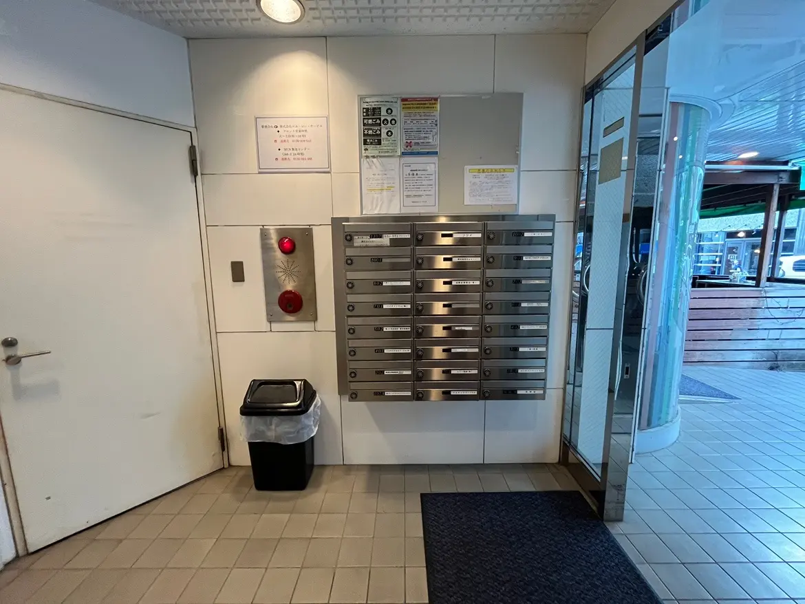 - 住宅のエントランスエリア、宅配ボックスの並ぶ壁面とゴミ箱、左側にドア、掲示板や警報装置が設置された内部の様子