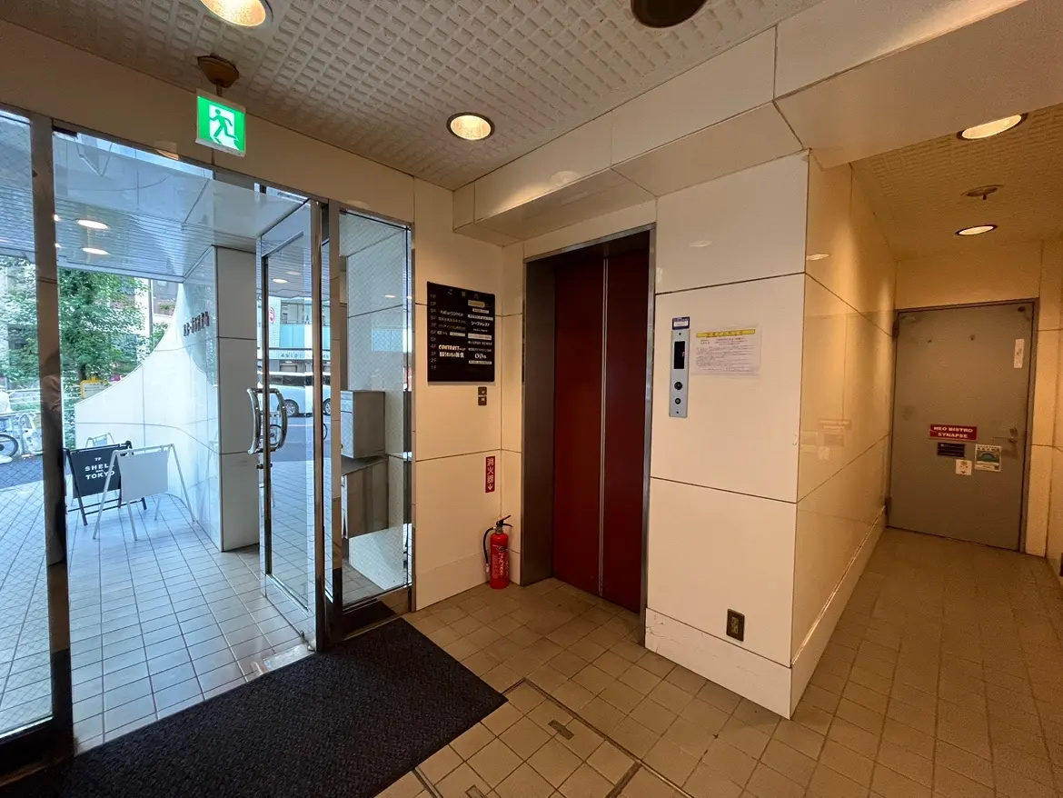 - ガラス扉のエントランス、エレベーターの横に案内板があり、外の自然光が差し込むモダンな建物のロビー