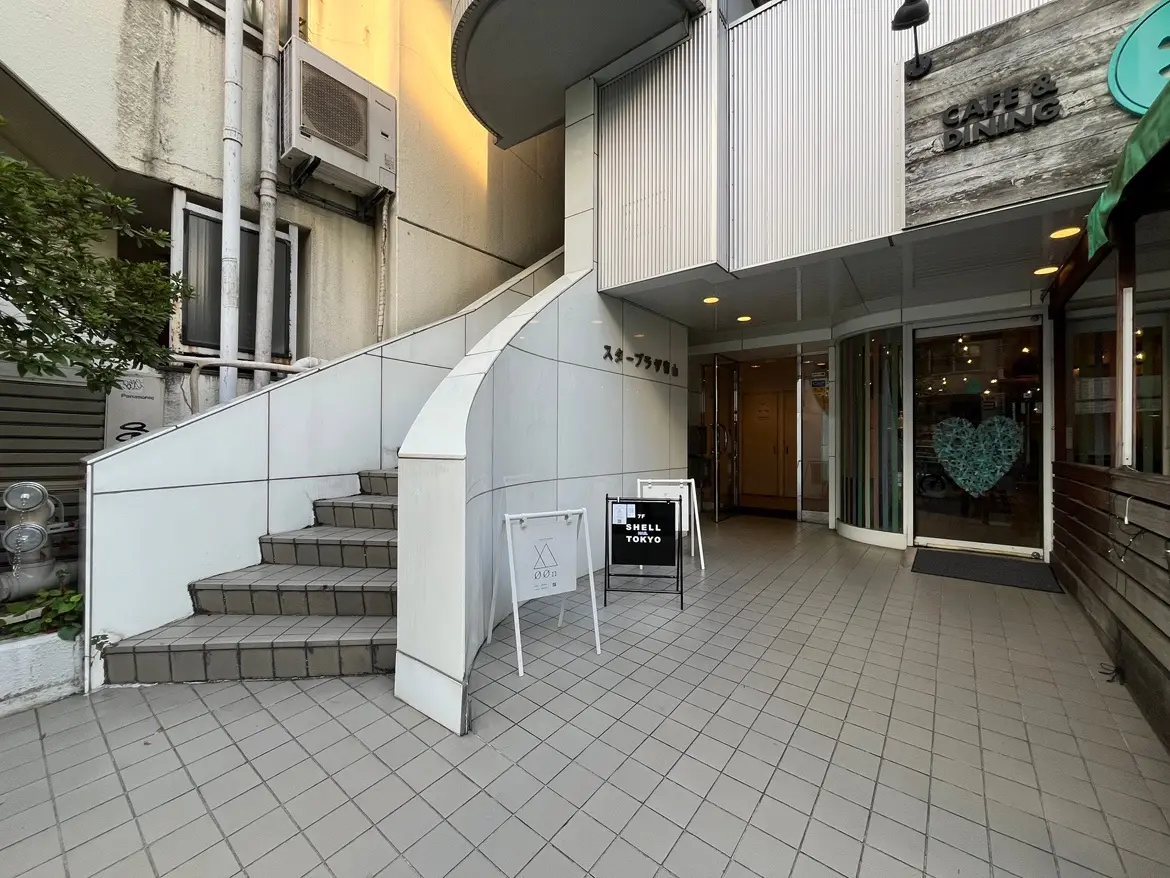 - スタイリッシュなカフェの入り口、白い階段とモダンなサインボードが特徴的、明るい照明とおしゃれなデザインの外観