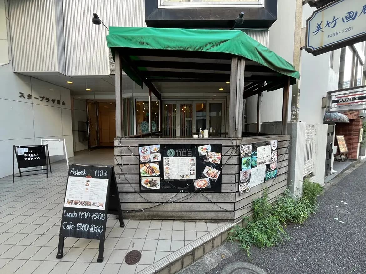 - 外観が木造のカフェ、緑のテントが特徴、メニューが掲示された看板と店舗前の歩道が映る街の風景