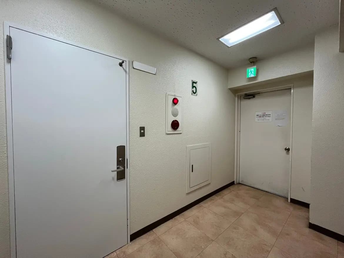 - 白いドアと緑の番号5が配置された廊下、壁には非常口とインターホンがあるシンプルなデザインの空間