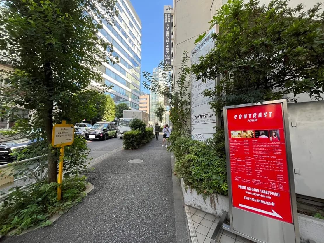 - 都会の通りに面した美容室「CONTRAST HAIR」の看板と周囲の緑豊かな街並み、商業ビルが立ち並ぶ風景