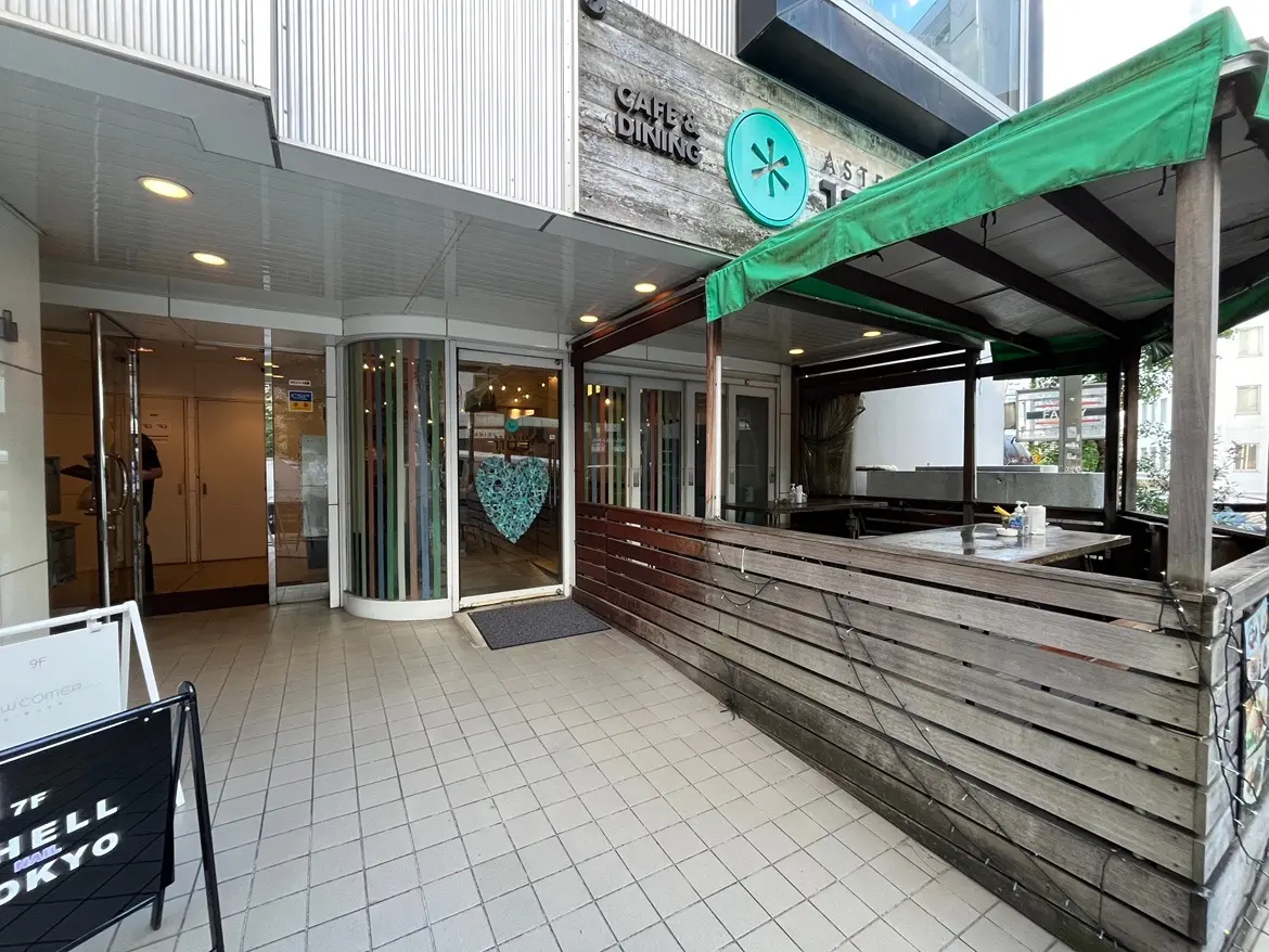 - カフェの入口とテラス席、木製の囲いと緑色の屋根が特徴的な外観、ハート型の装飾が施されたガラスの扉が見えるシーン