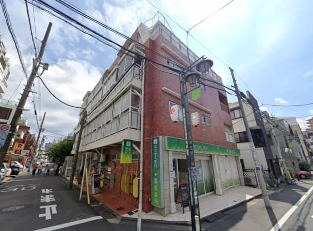 - 薄紅色のレンガ造りの建物、街角に位置し、通りの向こうに人々が歩く様子が見える風景、店先には自転車と看板が置かれている