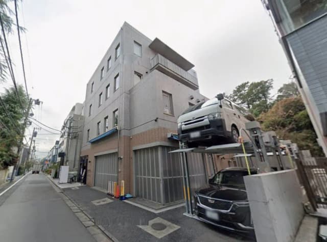 - 現代的なコンクリート造の建物と駐車場、上に立体駐車したバンと下の黒い車、狭い路地に面した外観