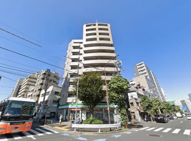 - 青空の下にそびえる高層マンション、近くにはファミリーマートやバス停があり、周囲は商業施設や住宅が立ち並ぶ賑やかな風景