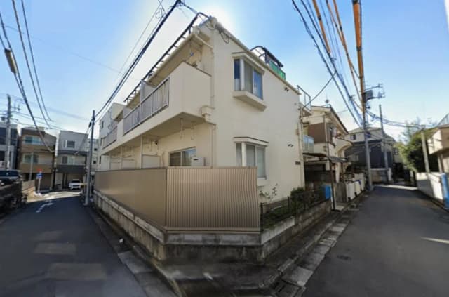 - 住宅街の角に位置する2階建てのアパート、周囲には電線と車道があり、外壁は淡い色彩のシンプルなデザイン