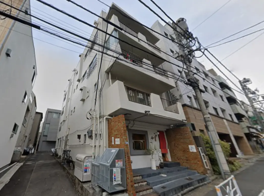 - 白い外壁が特徴のアパートの外観、洗練されたエントランスと階段、周囲に他のビルや電柱が立ち並ぶ風景