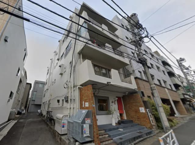 - 白い外壁が特徴のアパートの外観、洗練されたエントランスと階段、周囲に他のビルや電柱が立ち並ぶ風景