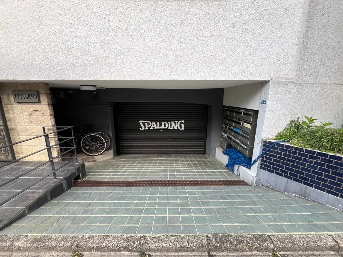- 駐輪スペースのある建物のエントランス、スラットシャッターに「SPALDING」のロゴ、タイル張りの床と郵便受けが並ぶ風景