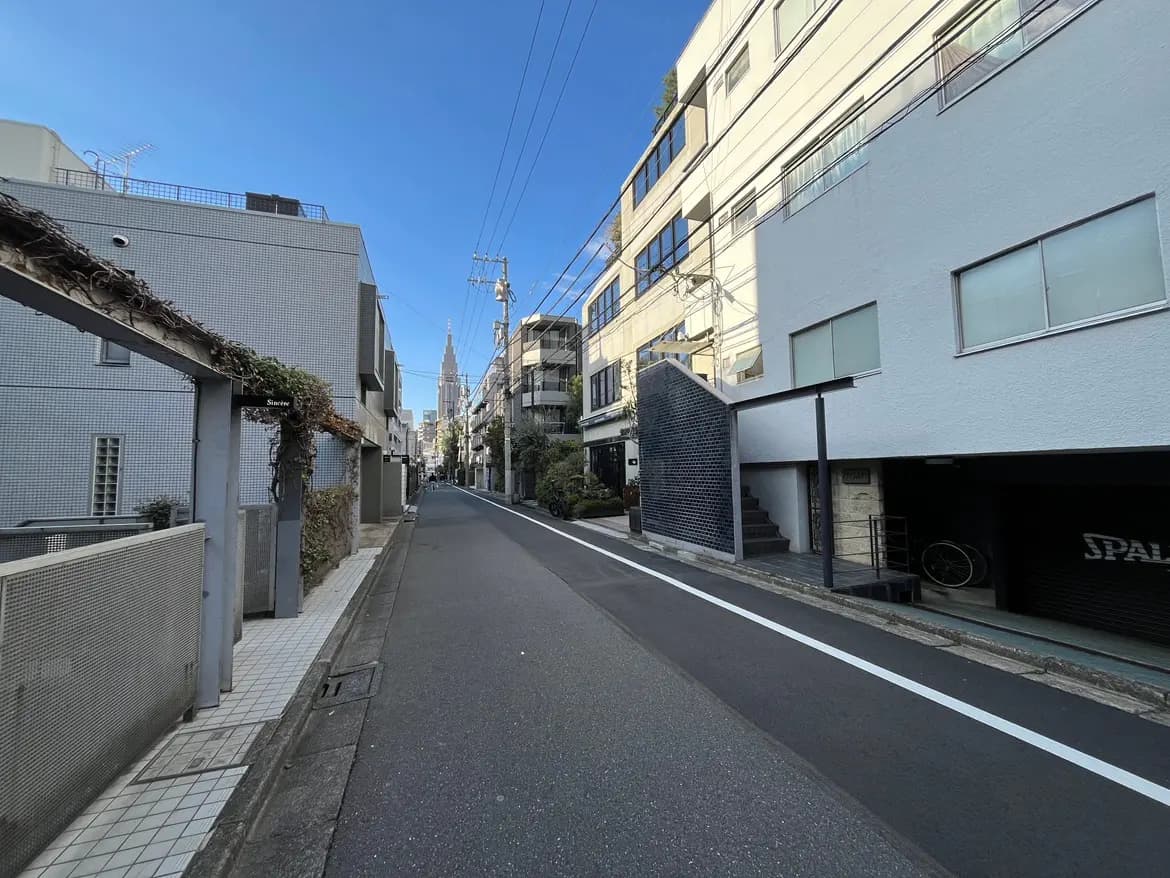 - 都市の静かな路地、賃貸マンションと住宅が並ぶ風景、遠くに見える高層ビルの尖塔が特徴的な青空の景色