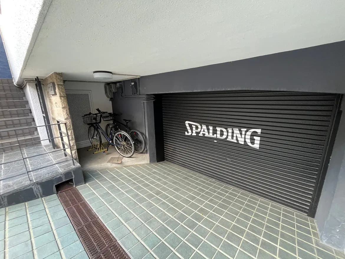 - 自転車が置かれたシャッター付きの駐輪場、モダンなデザインのエントランスと階段が見える景観