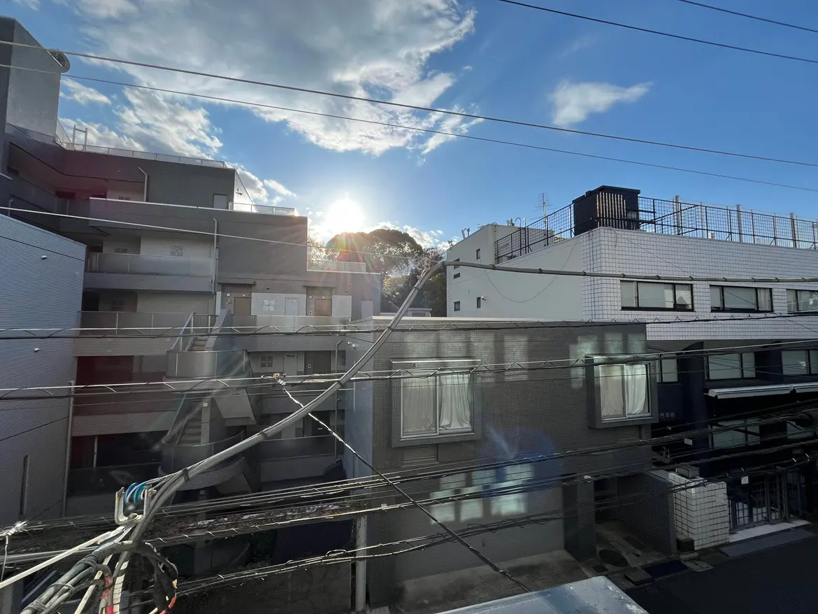 - 明るい青空の下に建つ住宅ブロック、手前の電線が交差し、背景に見える太陽が差し込む景観