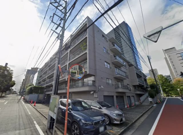 - 現代的なデザインのマンション外観、近くの道路と電柱、駐車中の車両、周囲に見える緑の風景