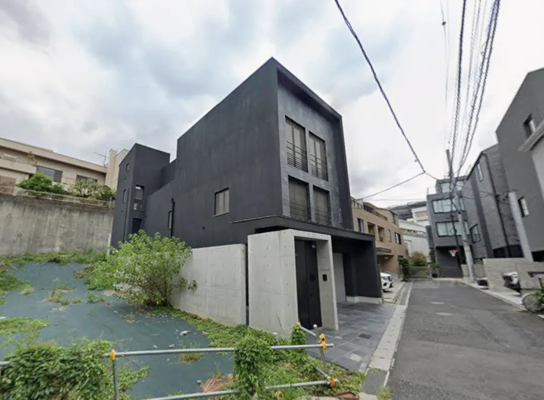 - モダンなデザインの黒い住宅、コンクリートの壁と大きな窓、周辺の緑と住宅街の風景が広がる外観