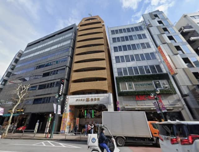 - 商業ビルが並ぶ街並み、中央に特徴的な外観のビル、手前には交通の往来がある繁華街の風景