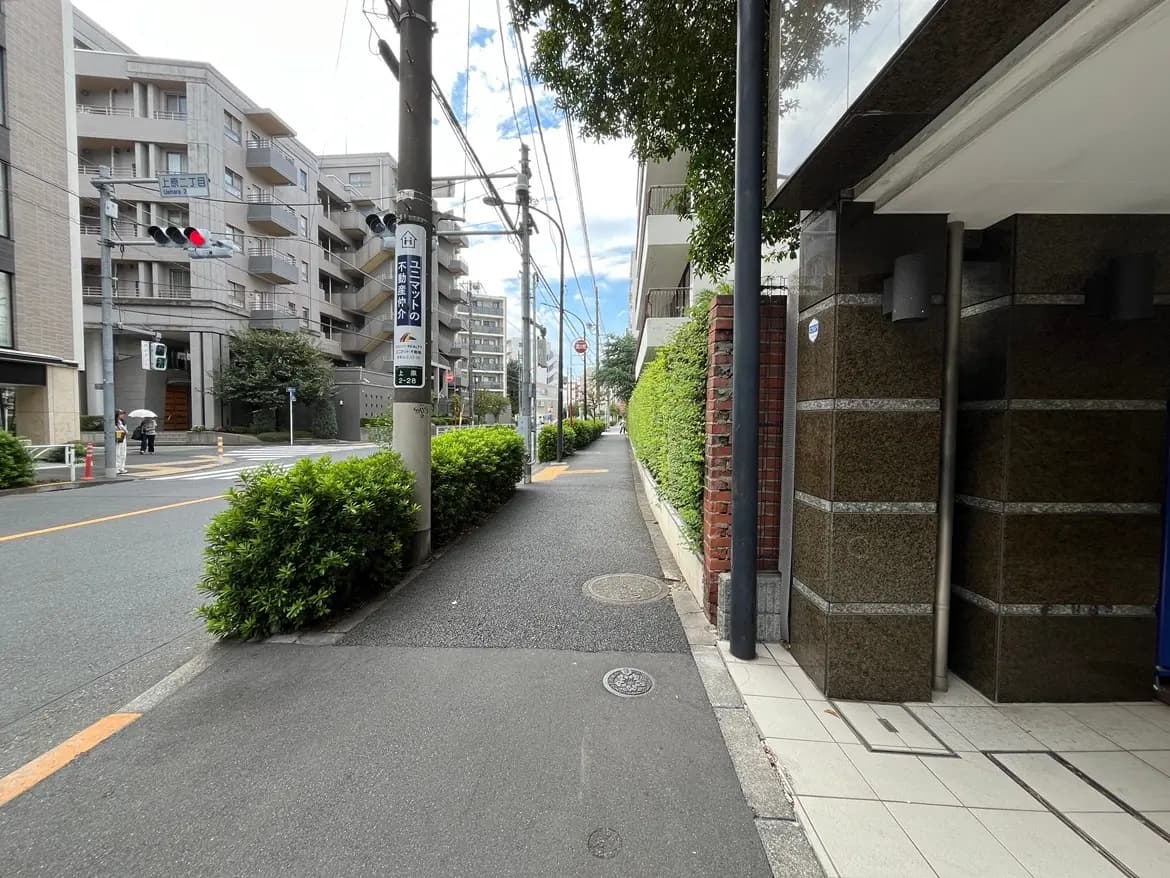 - 都市の街並みをイメージした歩道、両側に緑の植栽とマンションがある風景、青空と雲が広がる穏やかな日常の一コマ