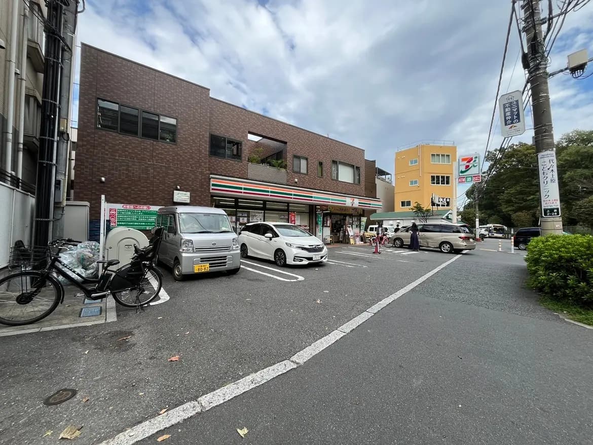 - セブンイレブンの店舗と駐車場、近くに自転車と車が停まっている街並み、曇り空の風景