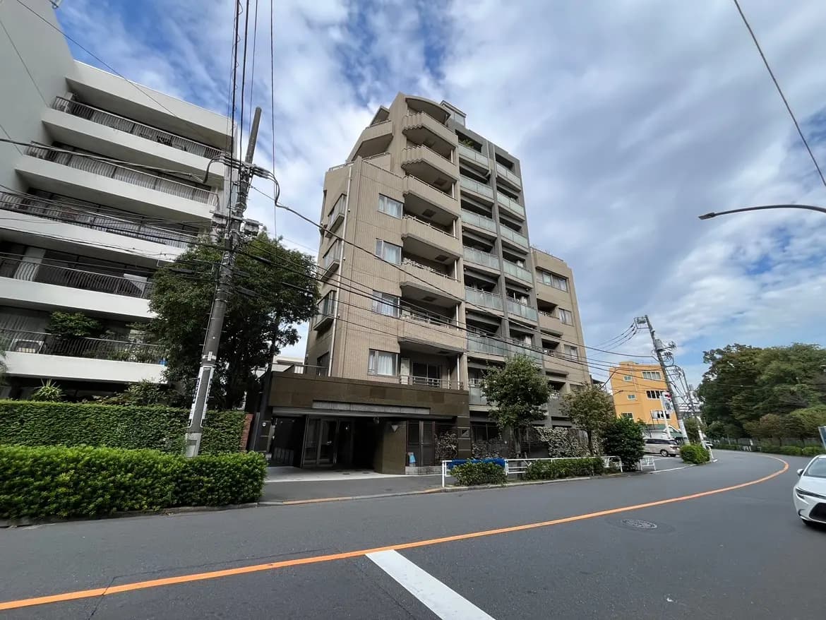 - 都市景観の中に建つマンション、周囲に緑がある、電柱や電線が見える通りの外観