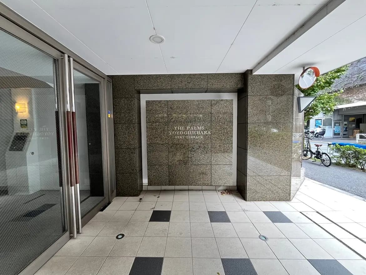- 洗練されたエントランスの外観、黒い石材の壁、フロアタイル、ロゴ「THE PALMS YOYOGIHARA」が中央にあるシンプルなデザインのエリア