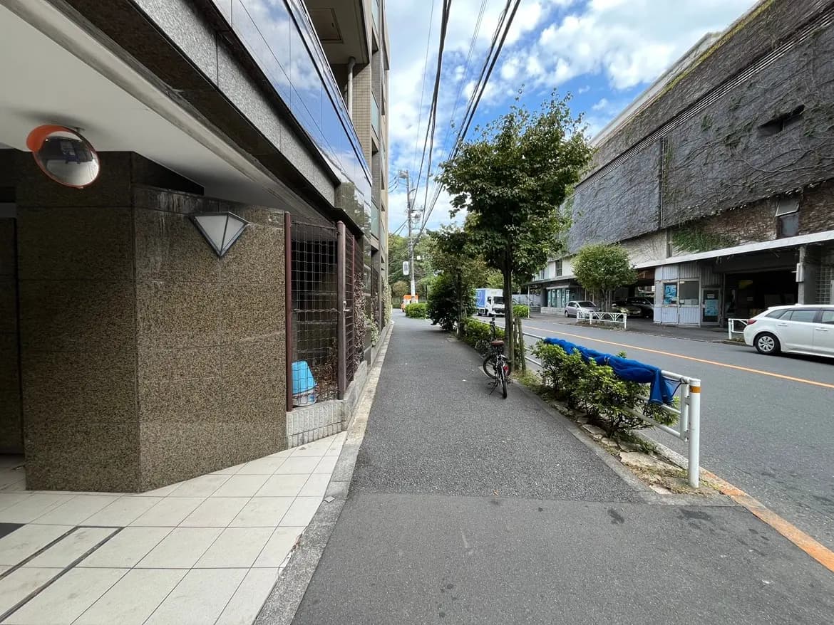 - 街中の歩道、左側にマンションの外壁と照明、右側に車道と自転車が停まる緑豊かな景観