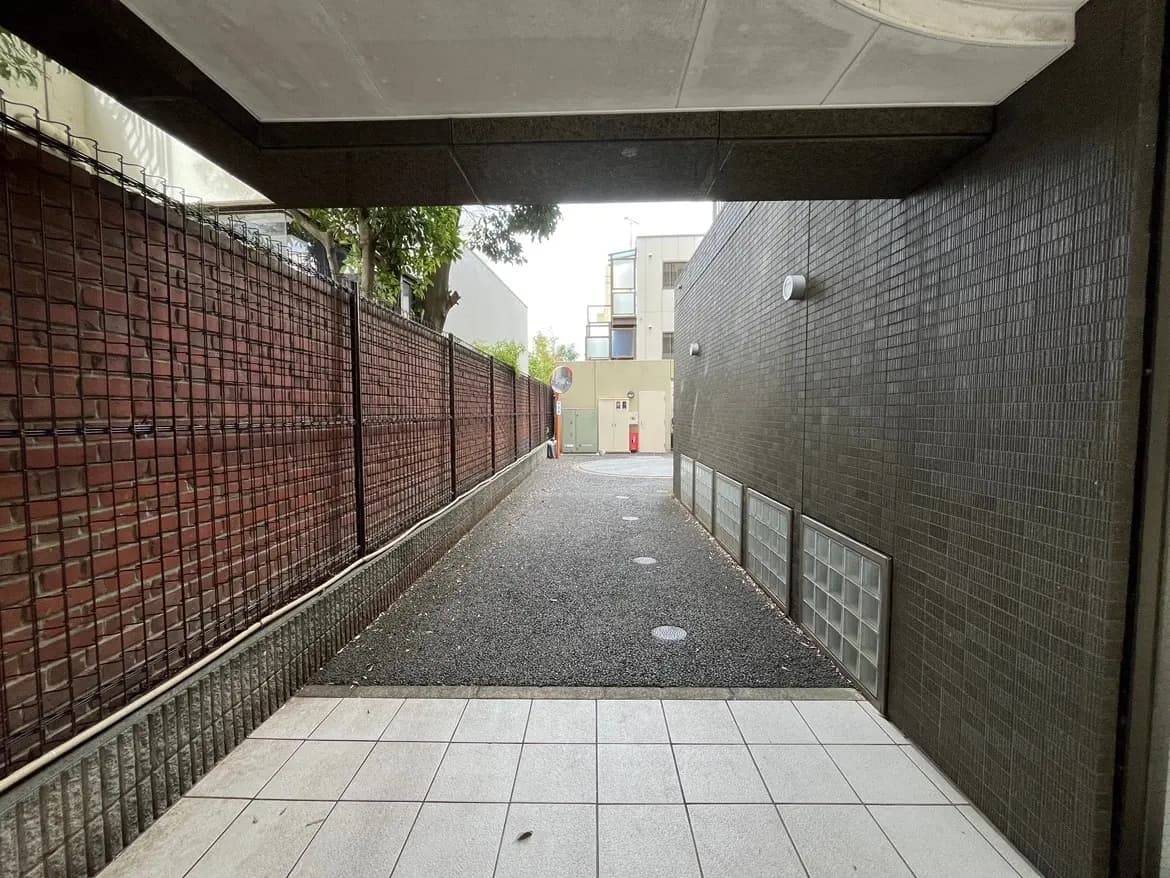 - 照明が当たる広いアプローチ、両側にレンガのフェンスがあるコンクリート製の通路、奥には建物の外観が見える賑やかな都市の一角