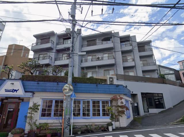 - 近代的なアパートメントビルとその前にあるカフェ、青い屋根と大きな窓が特徴の外観、周囲には緑が広がり、電線が交差する街並みの風景