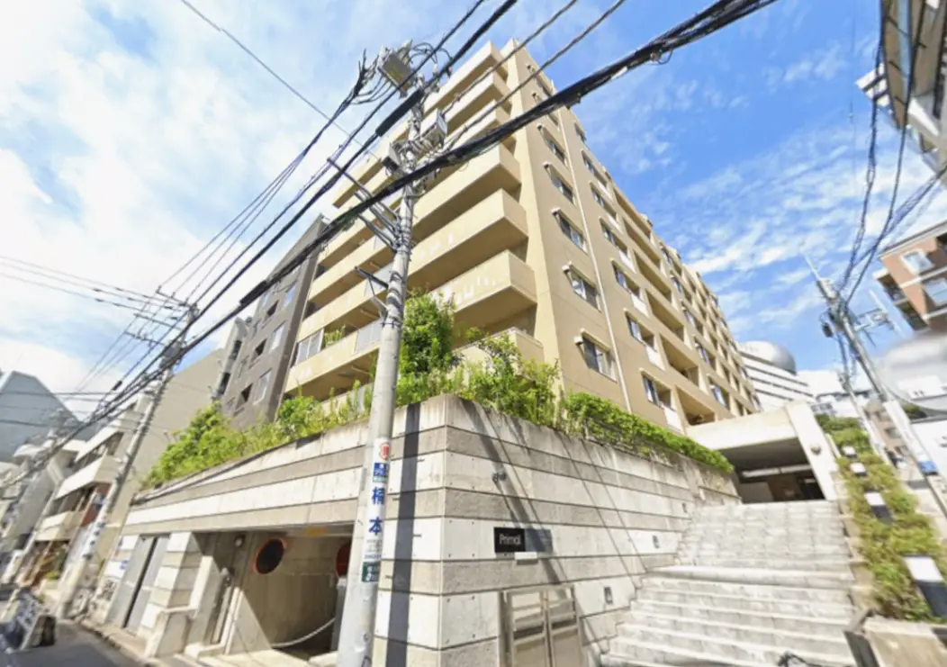- 都市型マンションの外観、明るい色合いの壁、緑の植栽とスロープ、周囲の電線が見える街並みの写真