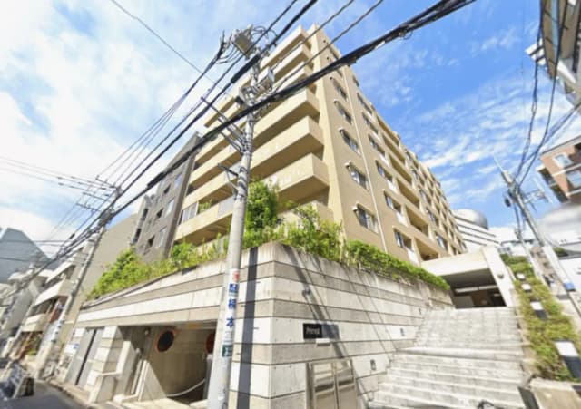- 都市型マンションの外観、明るい色合いの壁、緑の植栽とスロープ、周囲の電線が見える街並みの写真