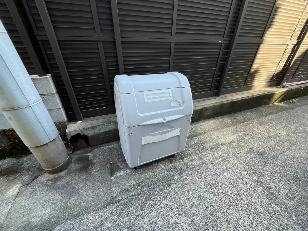 - 道路脇に置かれた白いゴミ箱、周囲はコンクリートの舗装、背景には黒いフェンスがある風景