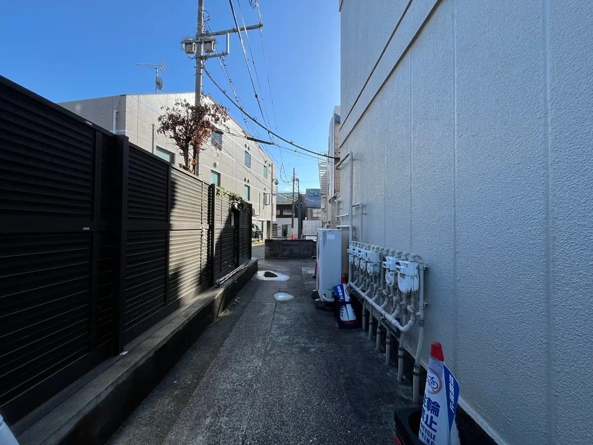 - 都市の住宅街の隙間、黒いフェンスと配管、青空が広がる狭い通路の風景