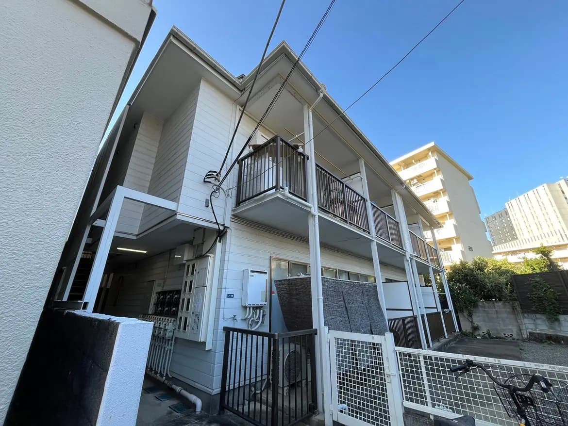 - 明るい空の下にある、木造の2階建てアパートの外観、バルコニーが特徴で周囲の建物とともに写っている全景